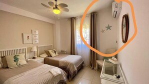 Une chambre d’hôtel avec deux lits, un ventilateur de plafond, une table de chevet avec une lampe, un miroir et une fenêtre avec des rideaux.