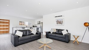 A modern living room with two black leather sofas, a glass coffee table, and a small side table. A kitchen with white cabinets and stainless steel appliances is visible in the background. - Scribbly Gum - Pet Friendly - 15 Mins Drive to Hyams Beach (Basin View)