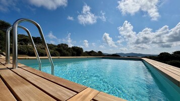 Piscina con bordo in legno e scaletta in metallo, circondata da alberi e montagne sotto un cielo sereno.