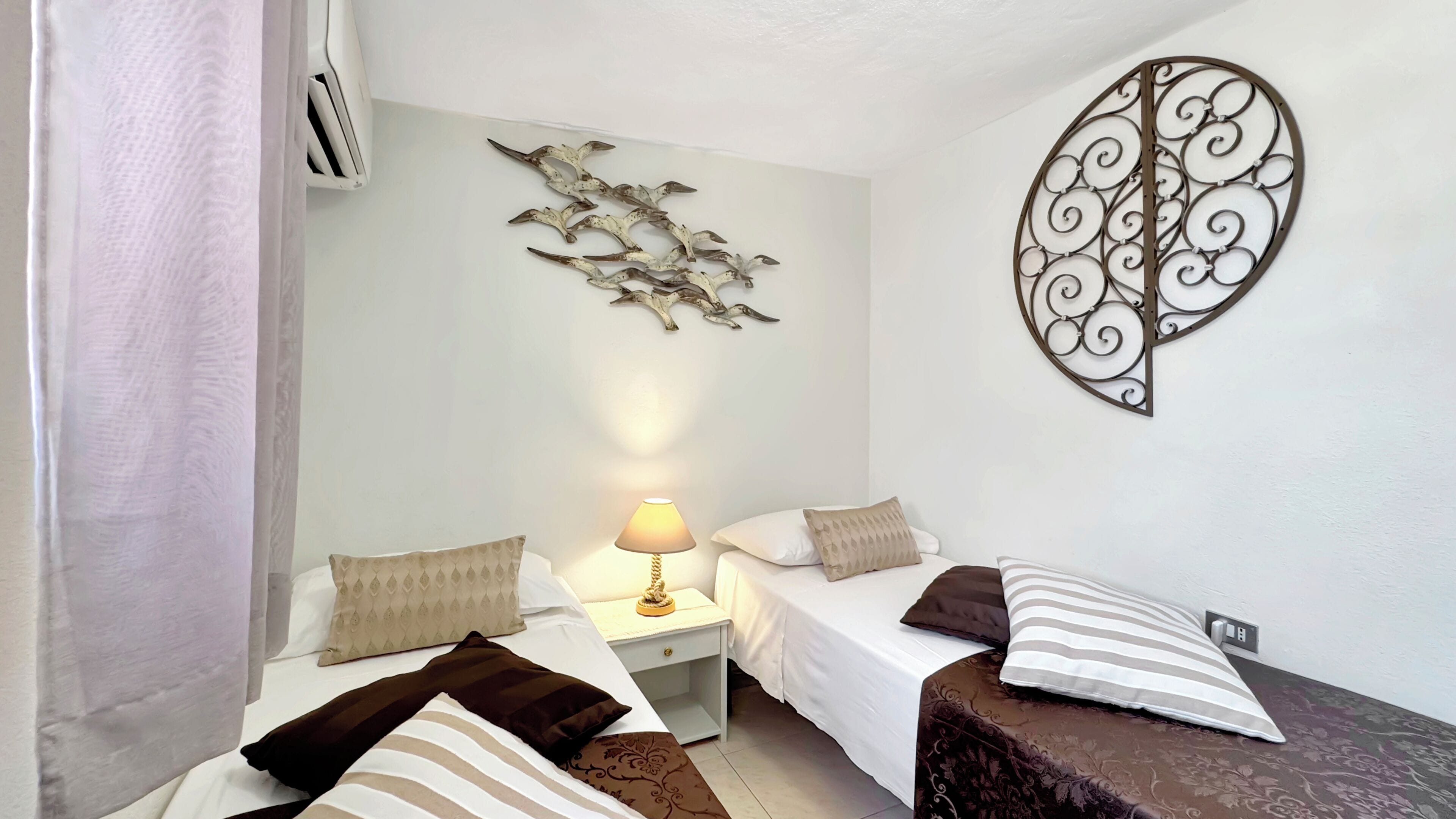 A hotel room with two beds, a nightstand with a lamp, decorative wall art, and a power outlet.