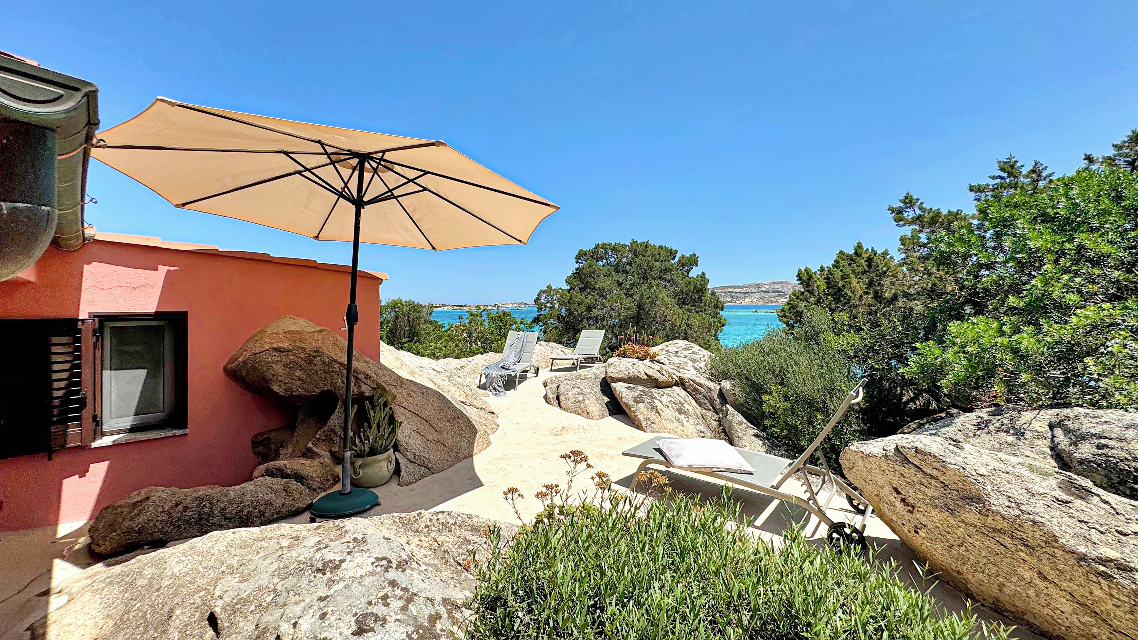 A patio with a beige umbrella, a chair, and a table, overlooking a scenic coastal view.