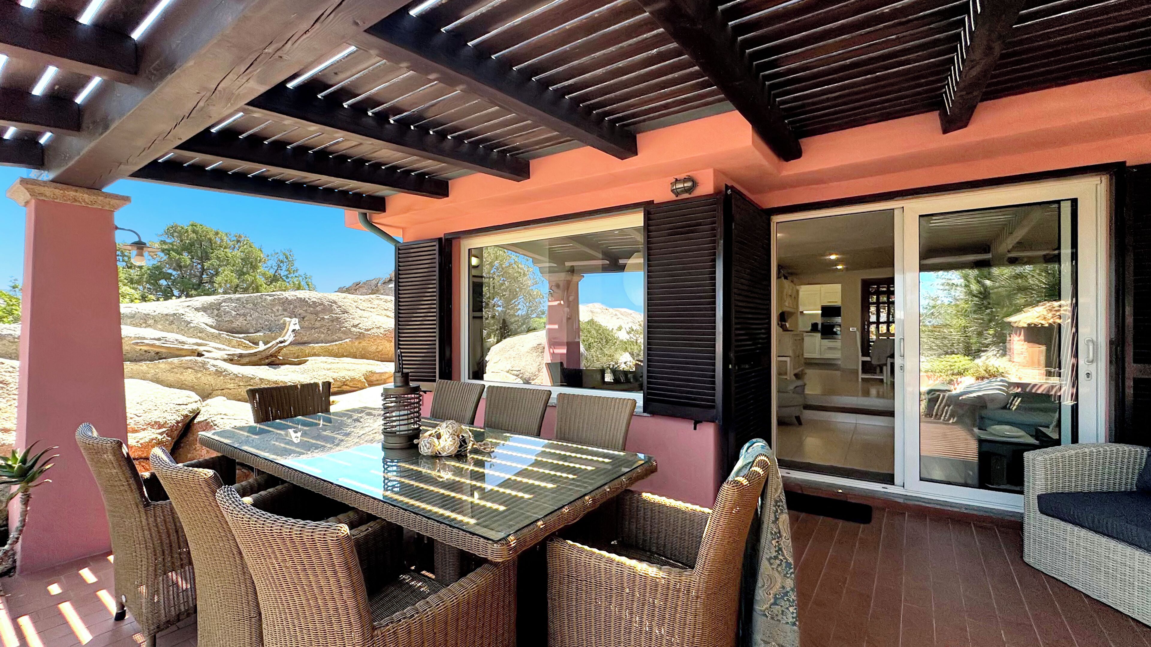 An outdoor patio with a glass table and wicker chairs, a pergola overhead, and a view of the natural landscape.
