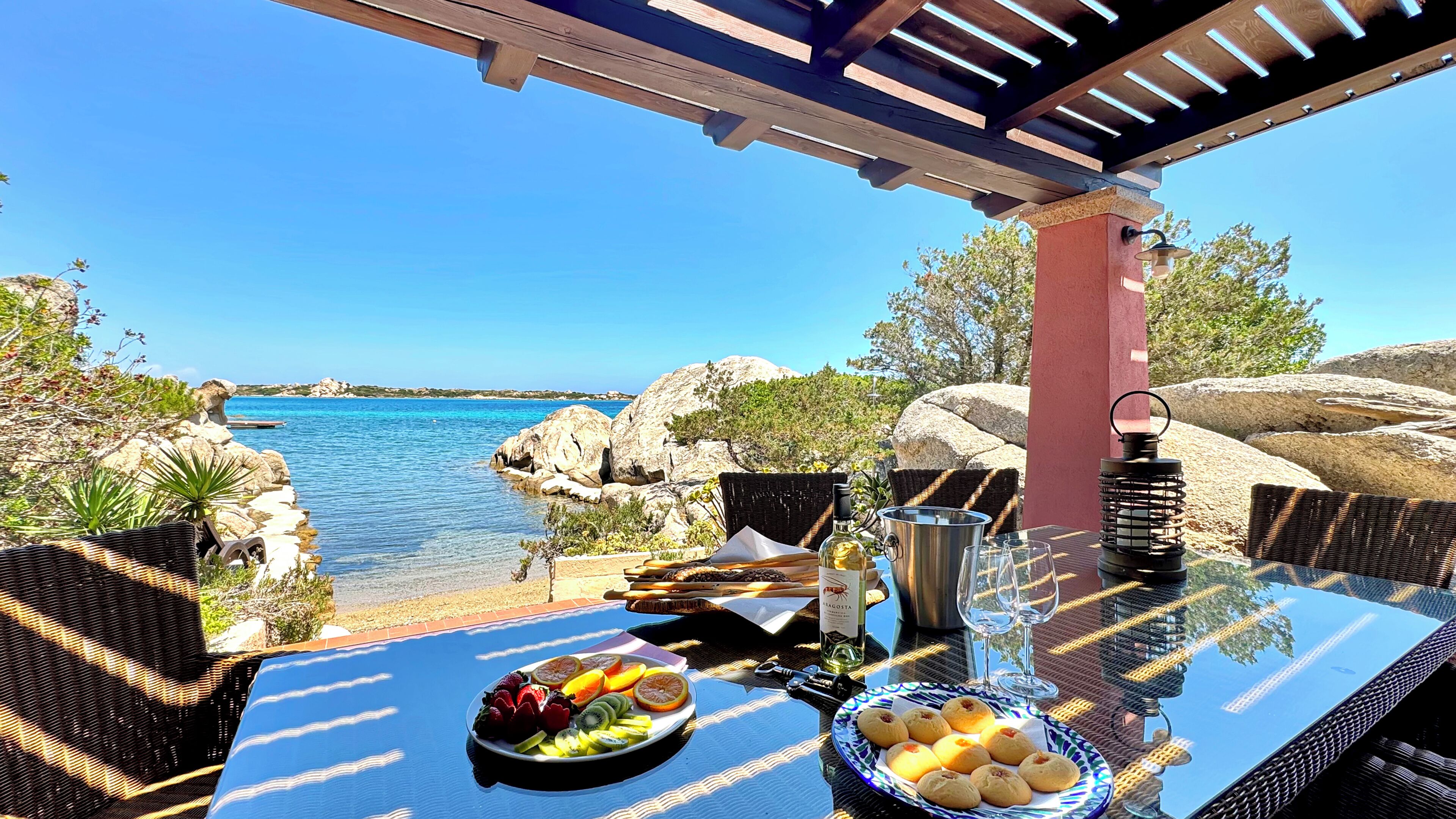 An outdoor dining area with a view of the sea, a table set with food and drinks, and a pergola overhead.