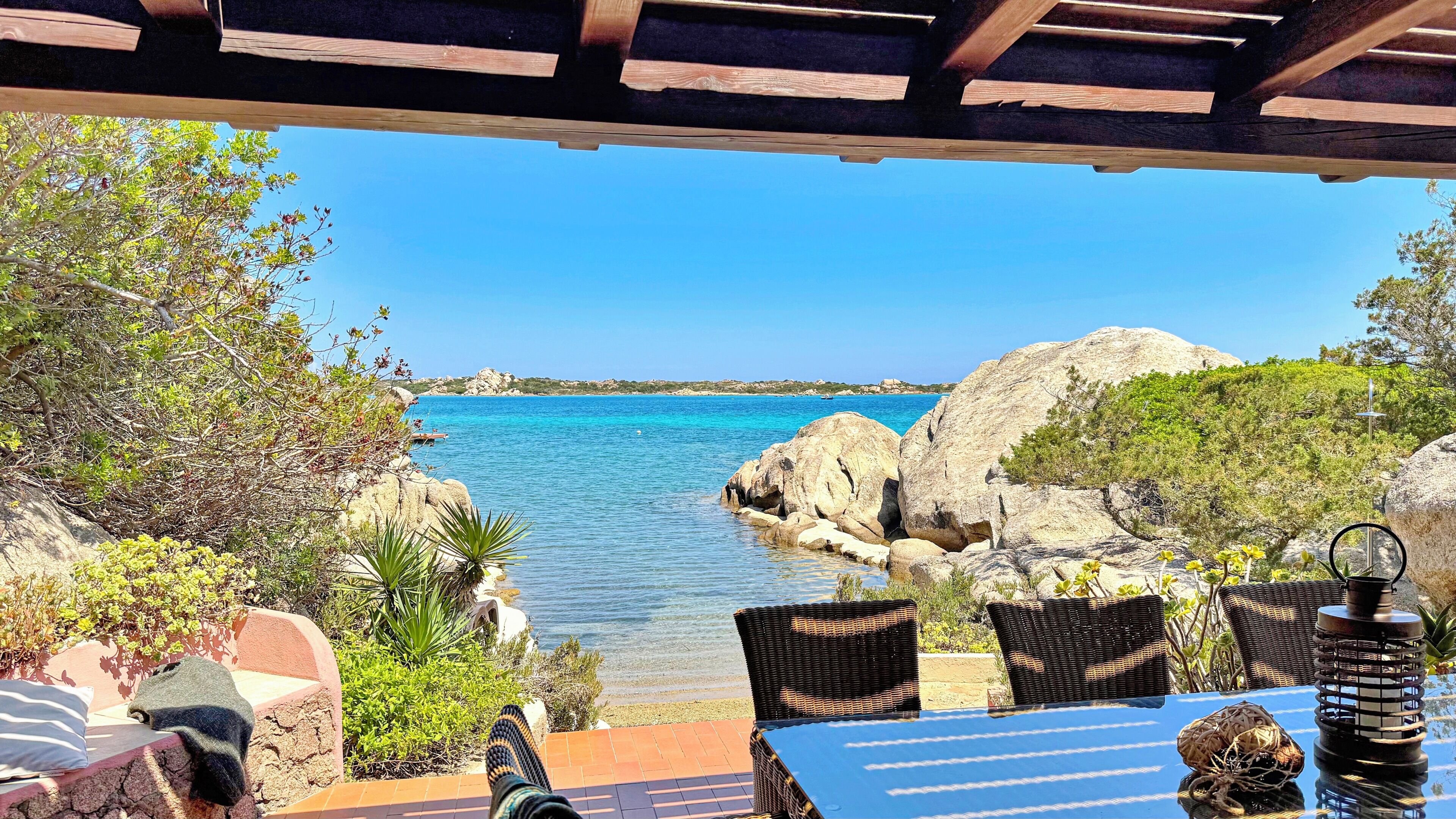 Un patio con sedie in vimini e un tavolo, con vista su una zona costiera caratterizzata da acque blu limpide e formazioni rocciose.