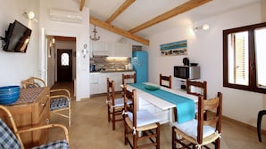 A dining area with a wooden table and chairs, a kitchen in the background, and a window with blinds. - Villa Iole (La Maddalena)