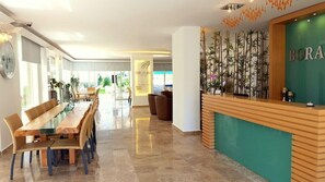 A hotel reception area with a wooden reception desk, a dining area with a long table and chairs, and a wall with vertical bamboo plants. - Bora Bora Apart Hotel (Alanya)