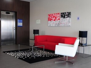 Un hall d’entrée moderne avec un canapé rouge, un fauteuil blanc, un tapis à motifs noir et blanc et deux lampes noires posées sur des pieds.