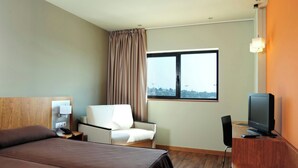 Habitación de hotel con cama, escritorio con televisor, una silla, ventana con cortinas y vista a la ciudad.