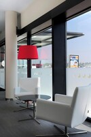 Un espacio de oficina moderno con un separador de vidrio, una mesa redonda, dos sillas blancas, una lámpara roja y vistas de la ciudad a través de amplios ventanales.