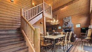 Interior de una cabaña de troncos con una escalera de madera, una zona de comedor con una mesa grande y sillas, una chimenea de piedra y un televisor.