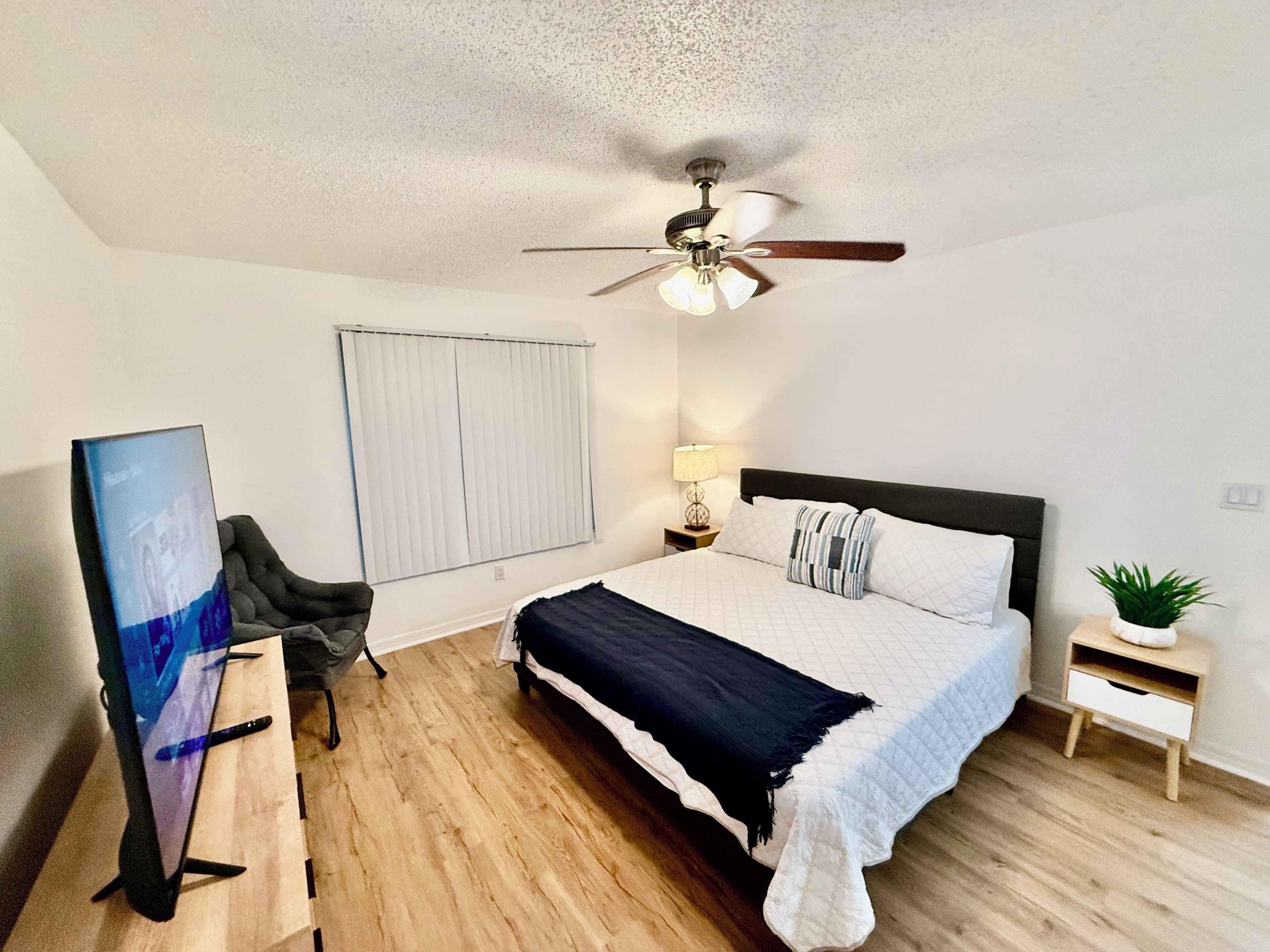 A bedroom with a bed, a ceiling fan, a window with blinds, a desk with a chair, and a small plant.