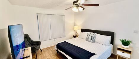 A bedroom with a bed, a ceiling fan, a window with blinds, a desk with a chair, and a small plant.