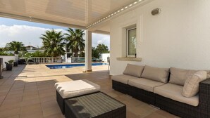 A covered outdoor area with a sofa and a coffee table, a pool, and palm trees in the background. - Stylish Villa for rent with 4 bedrooms and swimming pool on a wide channel in Empuriabrava (Empuria Brava)