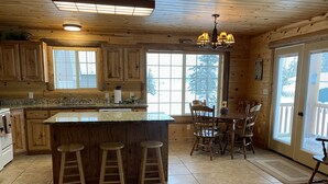 A wooden cabin-style kitchen with a central island, bar stools, a dining table with chairs, and large windows. - Comfortable cozy cabin, accessible to Snowmobile & ATV trails & Fly Fishing  (Island Park)