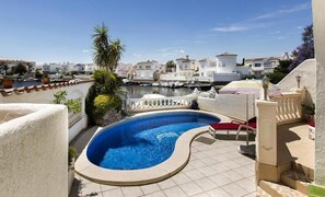 A rooftop swimming pool with a curved edge, surrounded by a tiled deck and a railing. In the background, there are white buildings and a marina with boats.