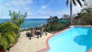 A swimming pool with a curved edge, surrounded by a stone patio. There are two lounge chairs and a green umbrella. The pool overlooks a coastal view with the ocean and a clear sky. - Montezuma Heights the cute Mariposa Cottage, into the great wide open.  Sleeps 2 (Montezuma)