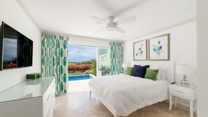 A bedroom with a bed, a TV, a ceiling fan, a nightstand, and a view of a pool.