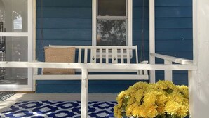 Un porche avec un banc blanc, un panier en osier et un vase rempli de fleurs jaunes.