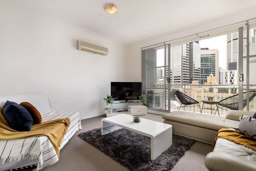 A modern living room with a white sofa, a white coffee table, and a TV. There is a large window with a city view.