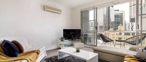 A modern living room with a white sofa, a white coffee table, and a TV. There is a large window with a city view.