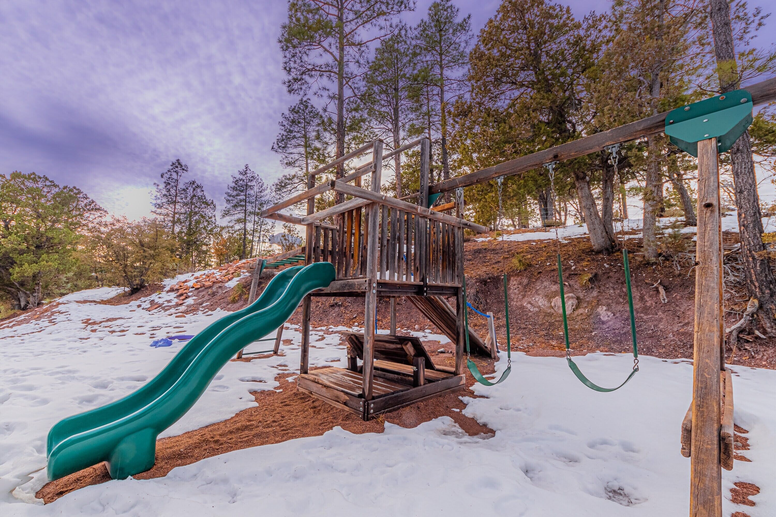 A wooden playground with a slide and swings, surrounded by trees and snow.