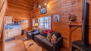A cozy cabin interior with wooden walls, a fireplace, a sofa, and a kitchen area.