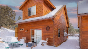 A wooden cabin with a snow-covered roof, a front porch with two Adirondack chairs, and a mailbox.