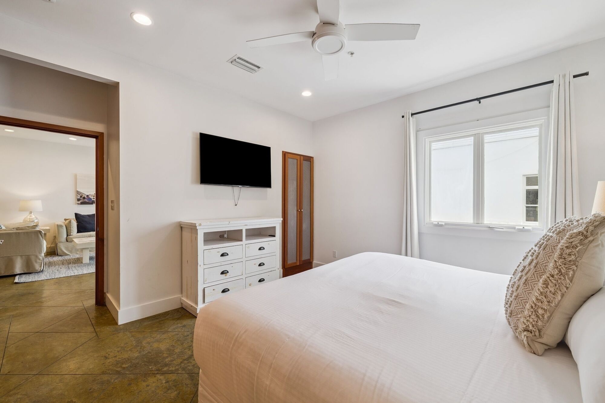 Une chambre à coucher avec un lit, un téléviseur fixé au mur, un ventilateur de plafond et une fenêtre avec des rideaux.