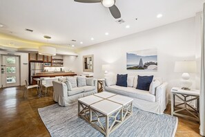 A modern living room with a sofa, coffee table, and lamps. There is a kitchen in the background with a refrigerator and cabinets.