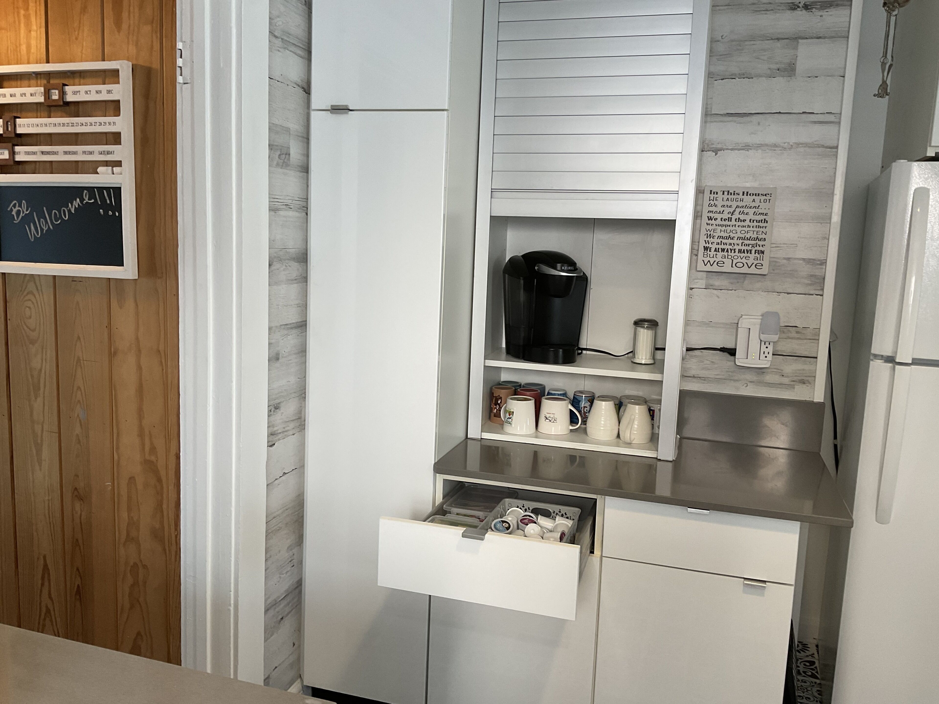 A kitchen with a coffee maker, mugs, and a calendar on the wall.