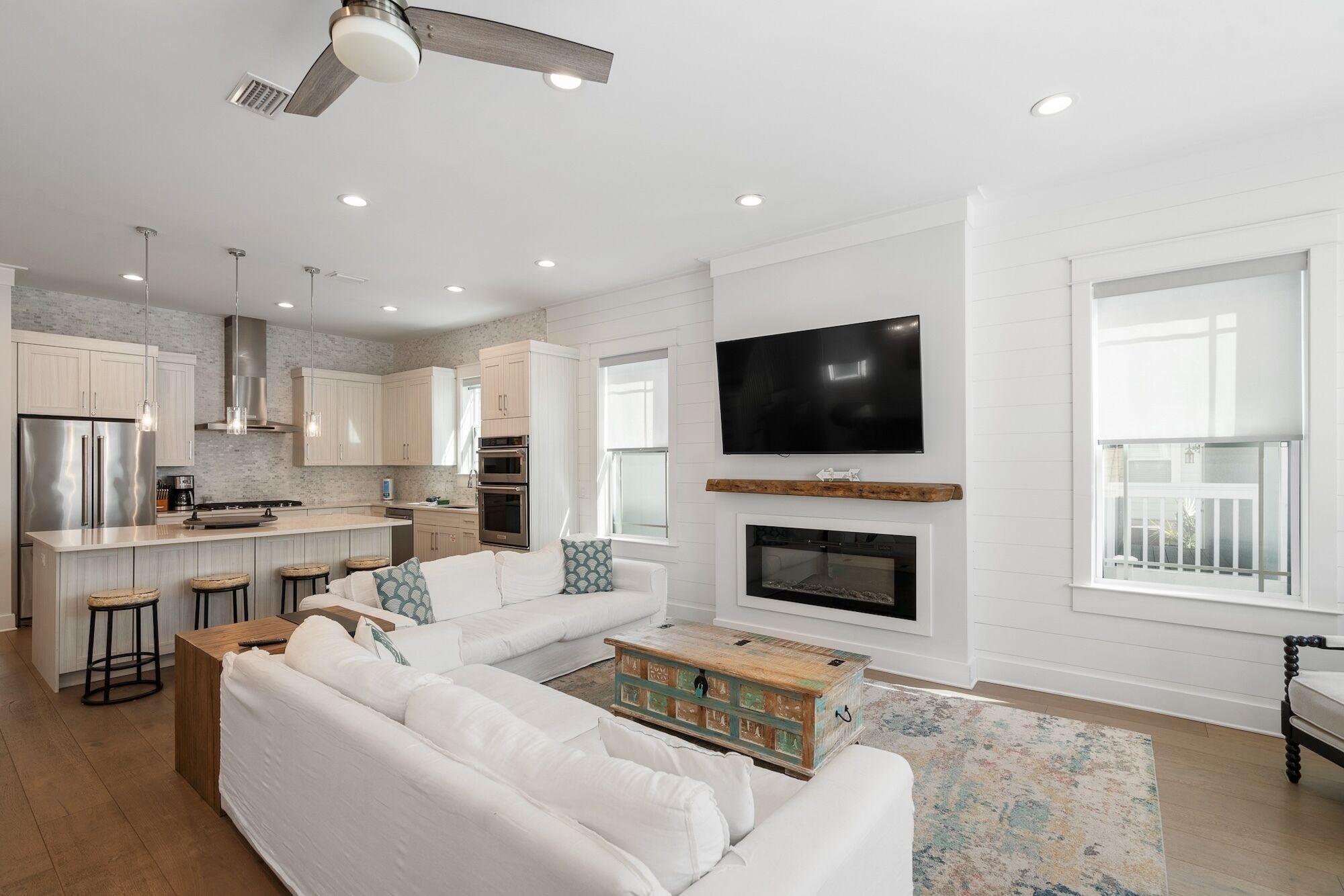 A modern living room with a white sofa, a coffee table, and a fireplace. There is a kitchen area with a refrigerator, oven, and sink. A TV is mounted on the wall above the fireplace.