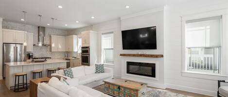 A modern living room with a white sofa, a coffee table, and a fireplace. There is a kitchen area with a refrigerator, oven, and sink. A TV is mounted on the wall above the fireplace.