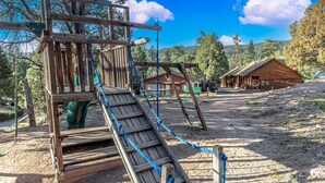 A wooden playground structure with a slide, surrounded by trees and a wooden building in the background. - Black Bear Cabin-onsite "bear hunt" & playground, game room, kitchen AC & heat (Payson)