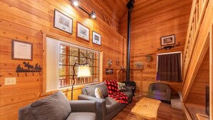 A cozy cabin interior with a sofa, armchair, and a small table. There is a window with a view of trees, a fireplace, and framed pictures on the walls. - Black Bear Cabin-onsite "bear hunt" & playground, game room, kitchen AC & heat (Payson)