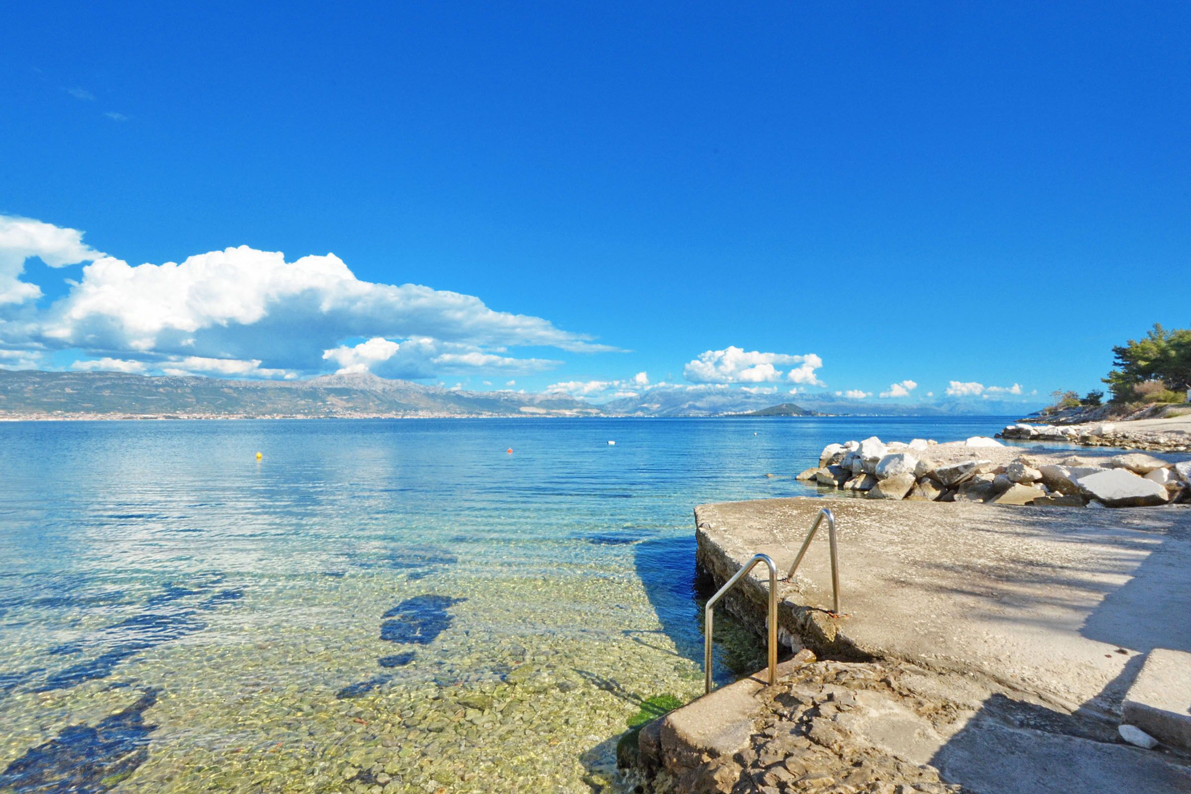 A clear view of a rocky shoreline with a metal staircase leading into the water, a calm sea, and distant mountains under a blue sky.