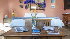 Un dormitorio con una cama, una mesita de noche de madera, una mesita junto a la cama con revistas y un libro de visitas, y un florero con flores.