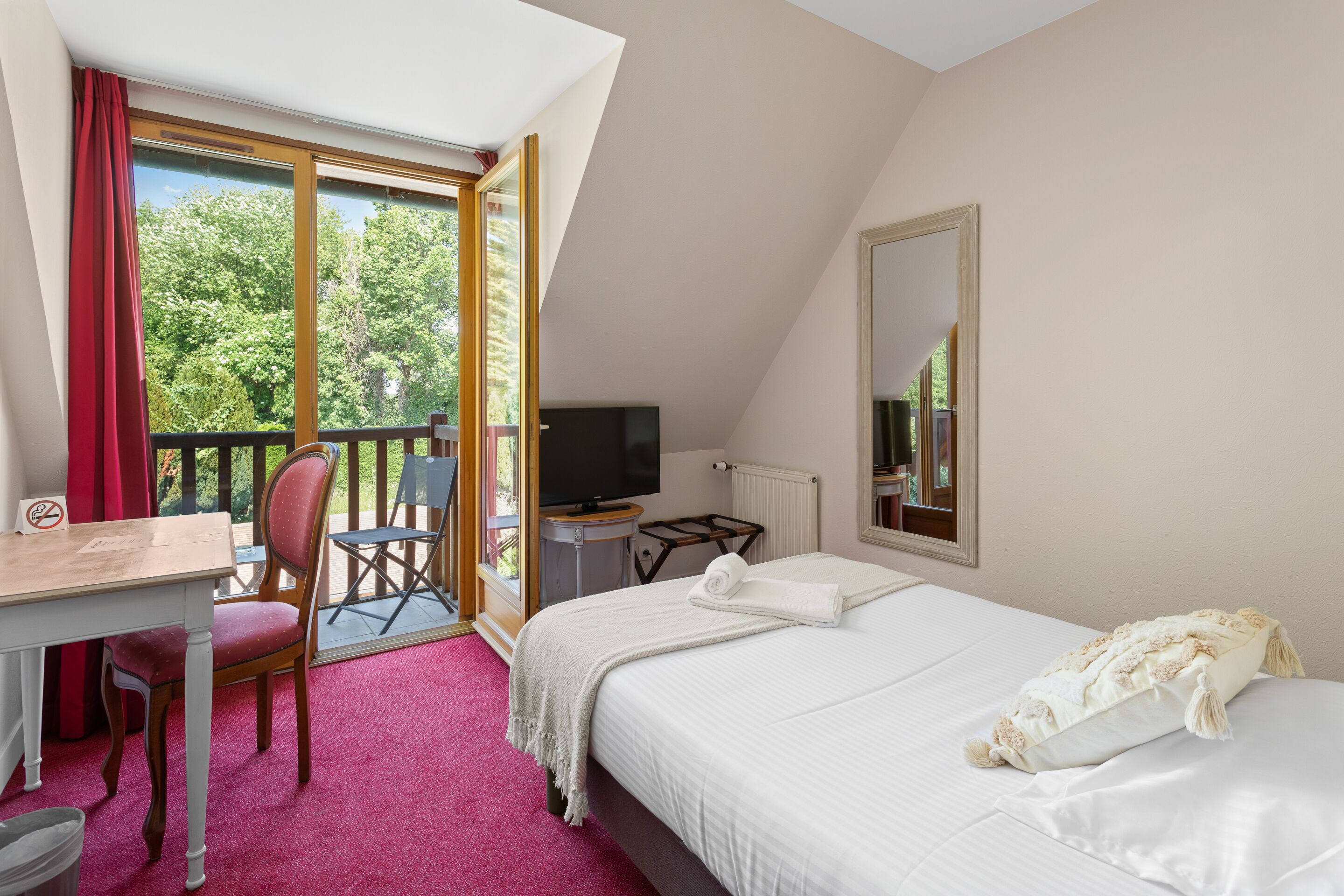 Quarto com cama, escrivaninha, cadeira, televisão e janela com vista para árvores.