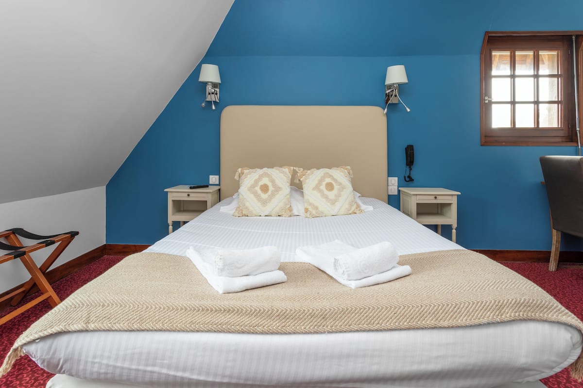 A neatly made bed with two pillows, a bedside table with a lamp, and a chair in a room with blue walls and a sloped ceiling.