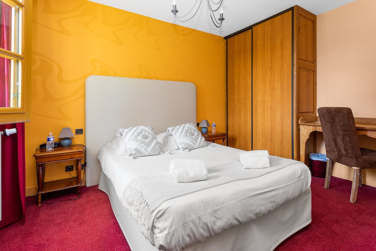 A hotel room with a large bed, a wooden wardrobe, a desk with a chair, and a red carpet.