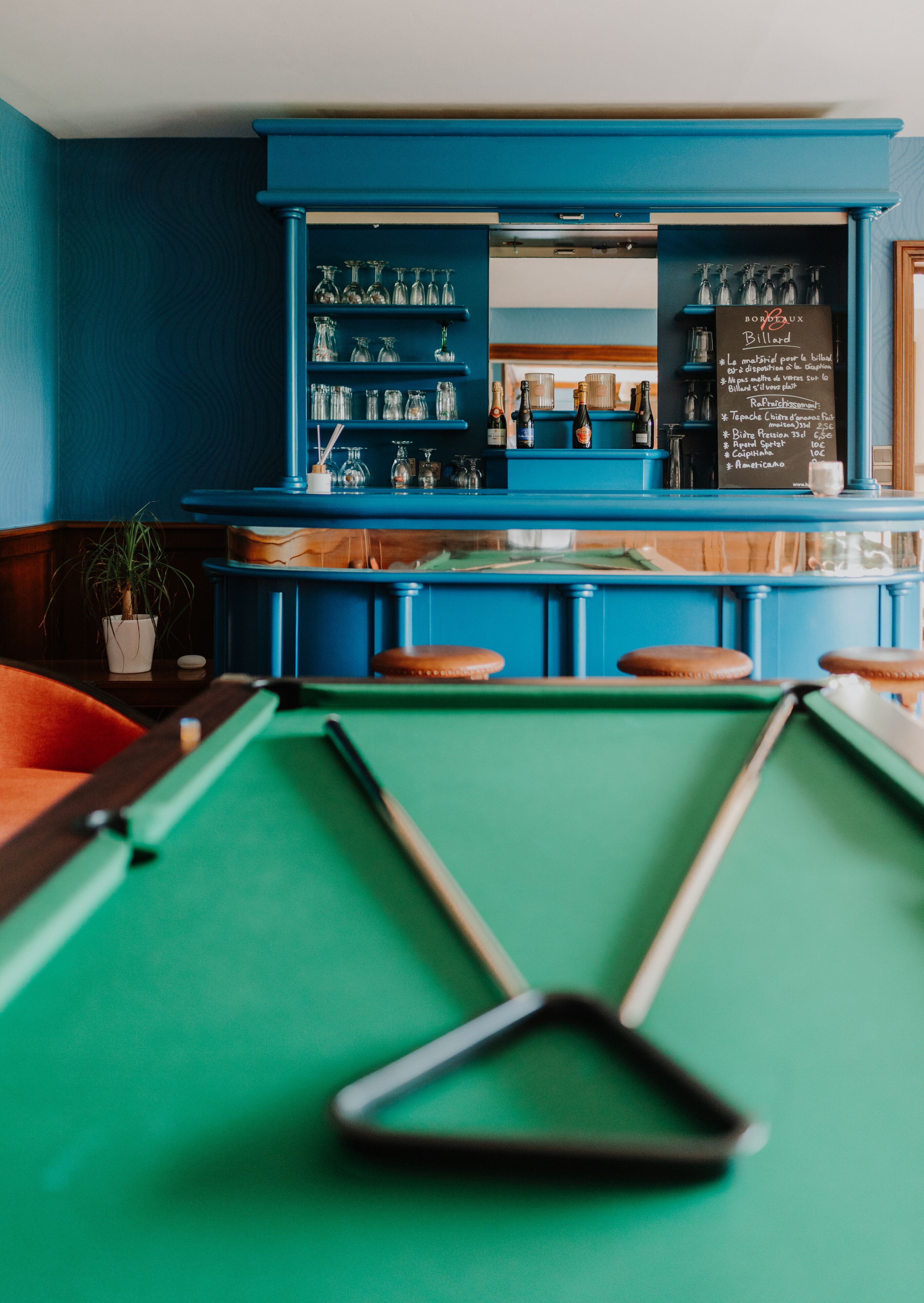 A pool table with a chalk and cue in the foreground, a bar area with shelves of bottles and glasses, and a chalkboard menu in the background.