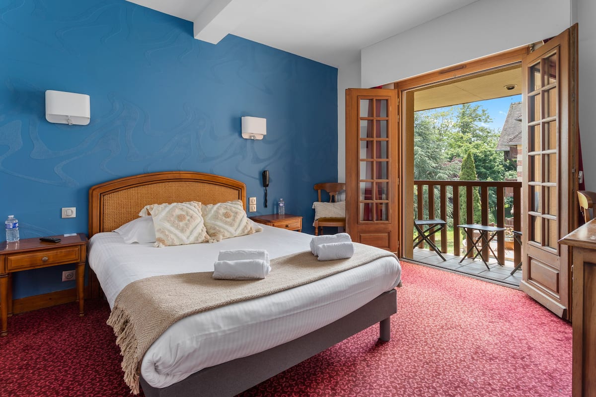 A hotel room with a large bed, a wooden headboard, a red carpet, two bedside tables with lamps, a wooden door leading to a balcony, and a view of greenery outside.