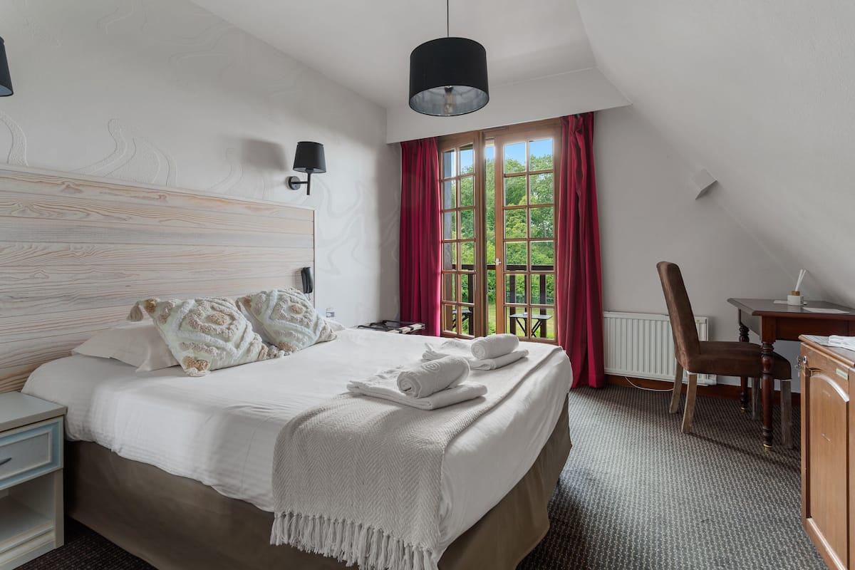 A neatly arranged bedroom with a bed, bedside table, lamp, and a view of the outdoors through a window.