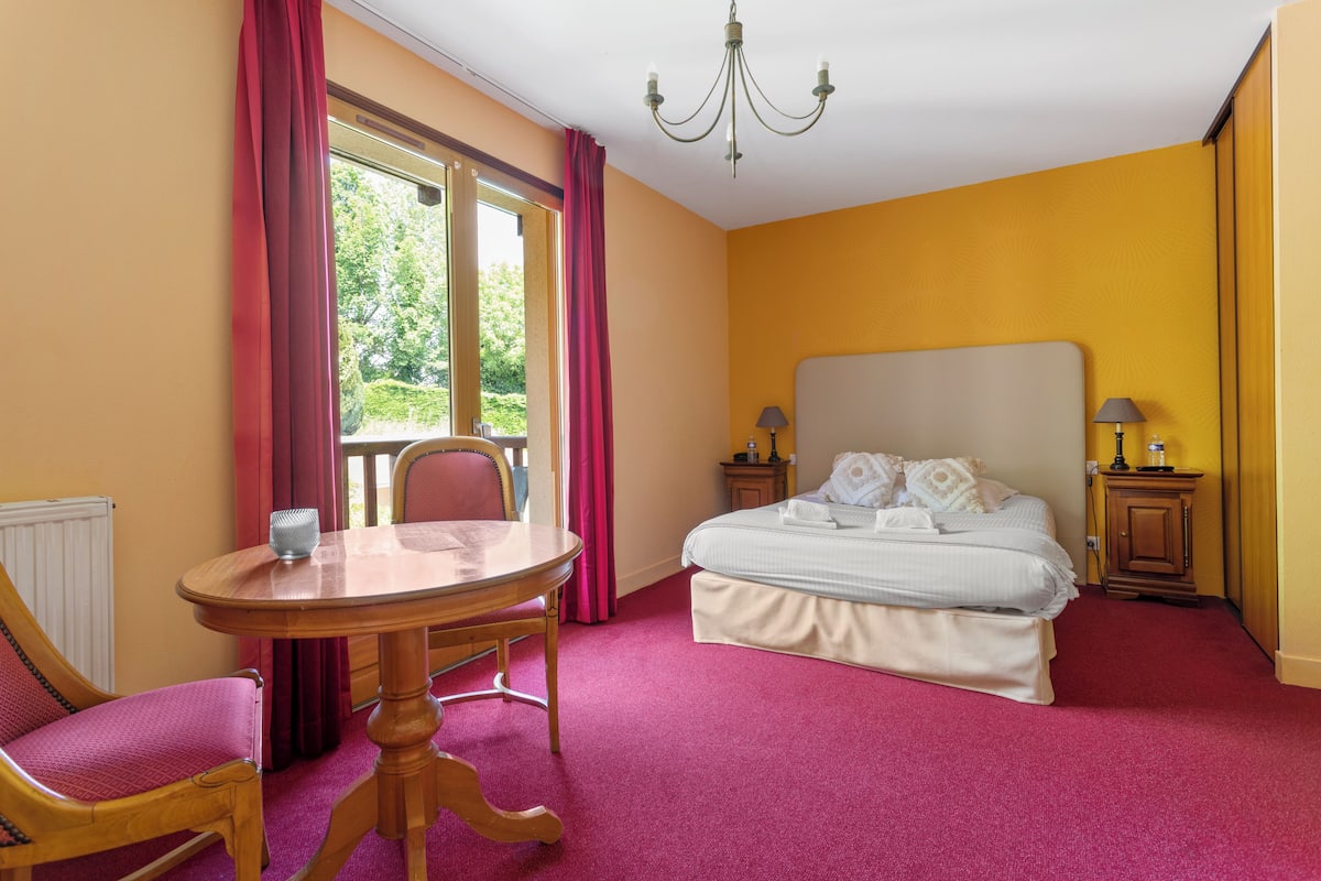 A bedroom with a bed, a wooden table, a chair, a window with curtains, and a chandelier.