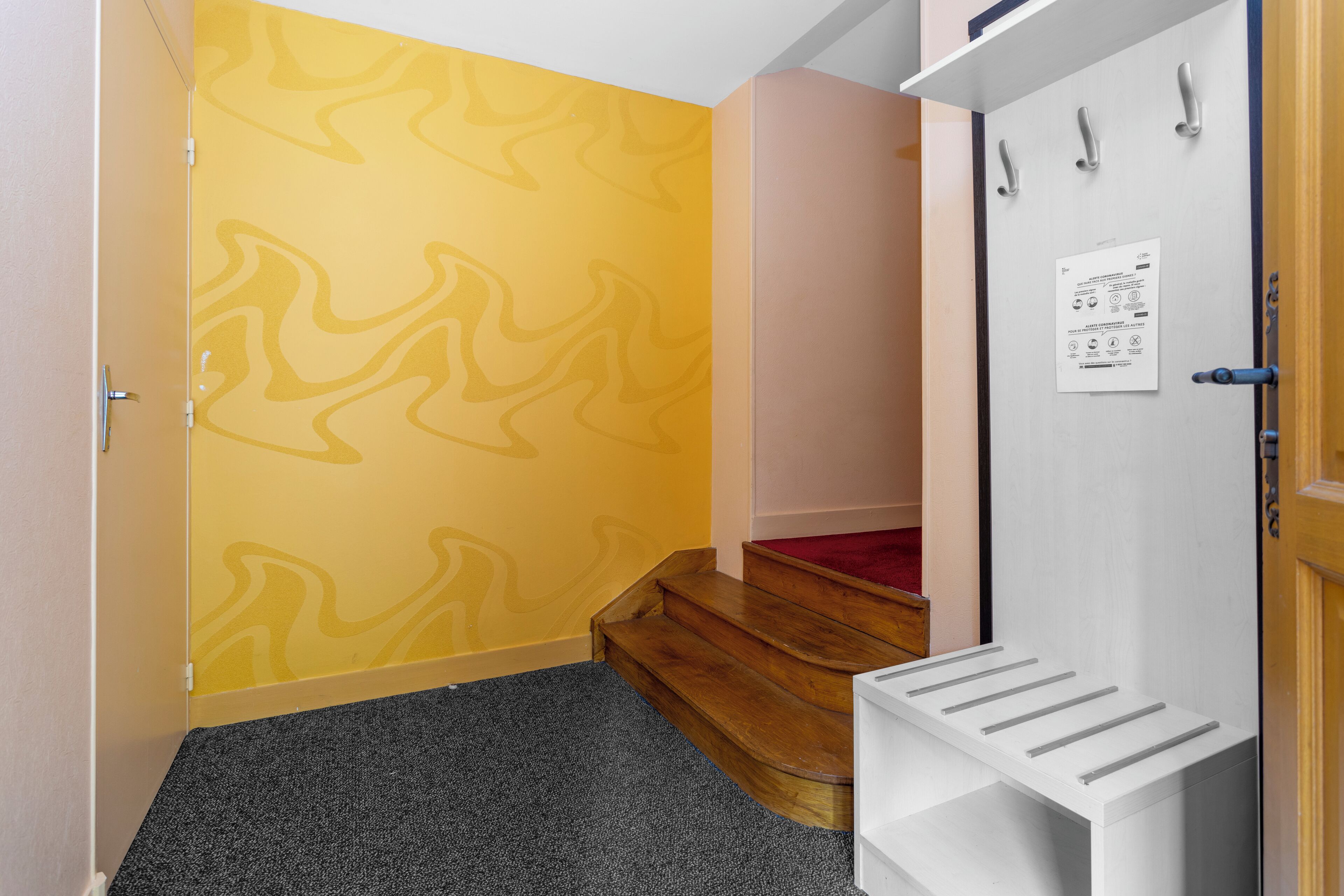 A hallway with a patterned yellow wall, a wooden staircase, and a white door with a notice.