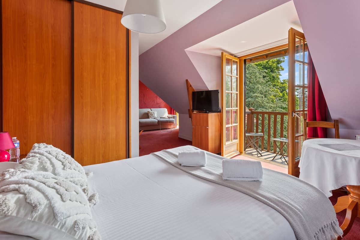 A bedroom with a large bed, a TV, and a balcony with a view of trees.