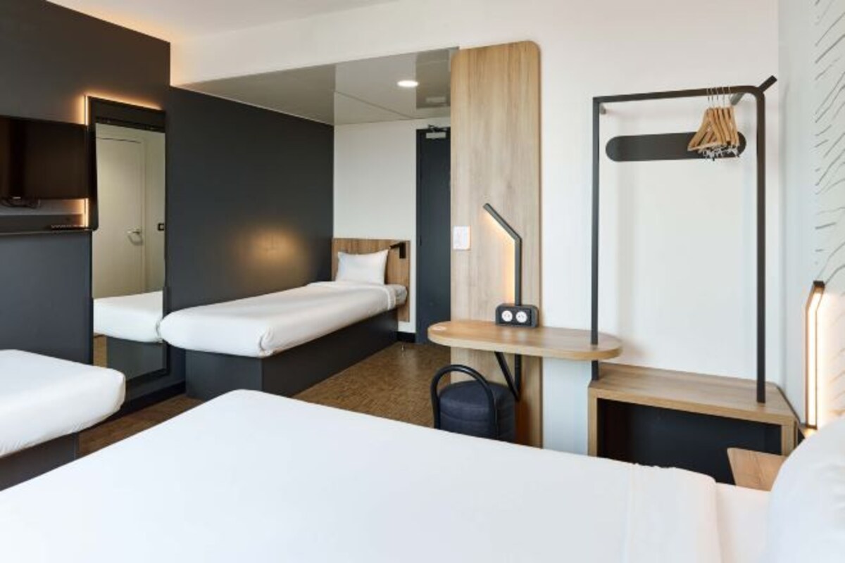 Une chambre d’hôtel moderne avec deux lits simples, une petite table avec une lampe, un porte-manteau et un téléviseur fixé au mur.