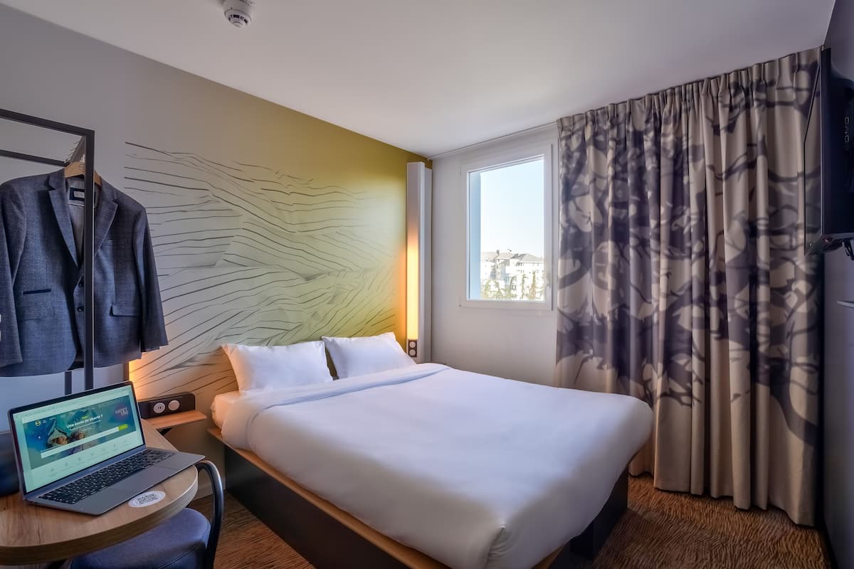 A hotel room with a bed, a laptop on a side table, a coat rack, and a view of buildings through the window.
