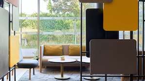 A modern lounge area with a sofa, two round tables, and a large window offering a view of greenery. - B&B HOTEL Paris Créteil (Creteil)