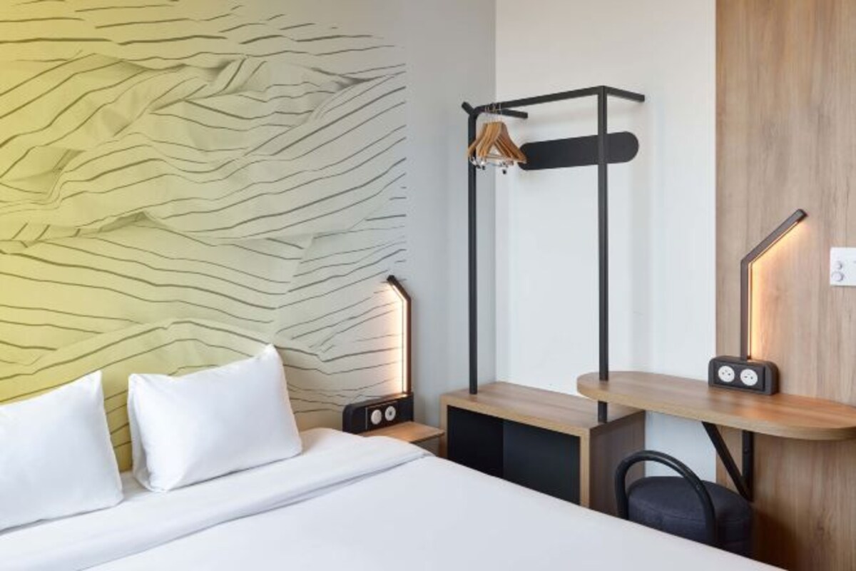 Une chambre d’hôtel moderne, équipée d’un lit, d’une table de chevet avec une lampe, d’une petite table en bois et d’un porte-manteau fixé au mur.