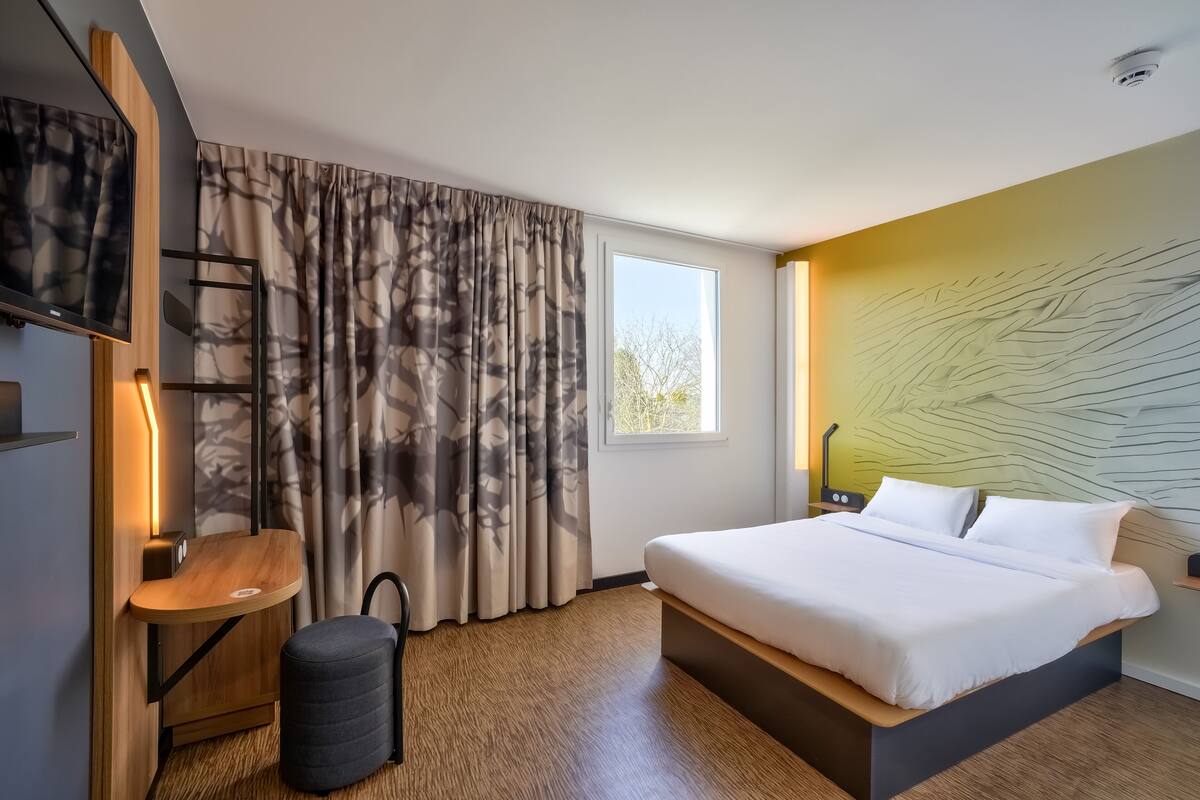 A modern hotel room with a bed, a desk, a chair, and a large window.